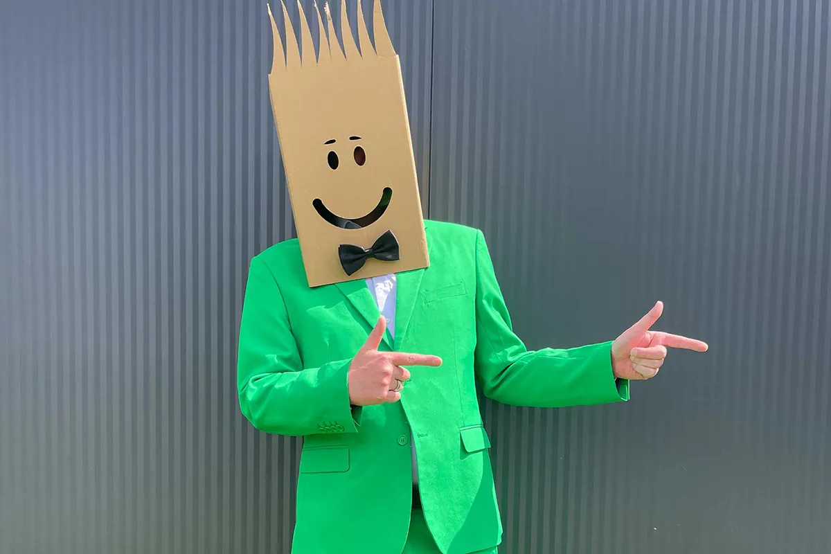 Herr Packners mit Kartonkopf und grünem Anzug steht vor einer Wand und zeigt mit beiden Händen zur Seite