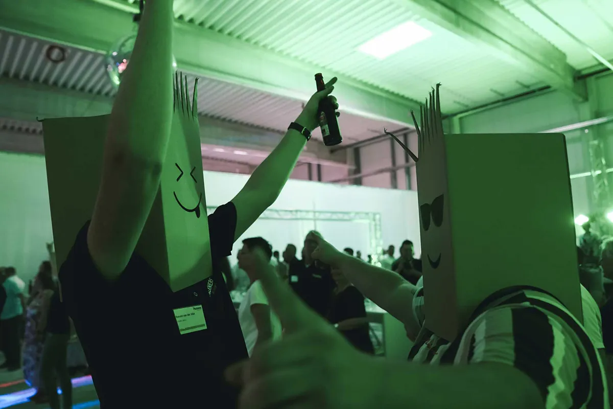 Zwei Personen mit Kartonkopf-Kostümen feiern auf einer Veranstaltung, eine hebt eine Flasche in die Luft