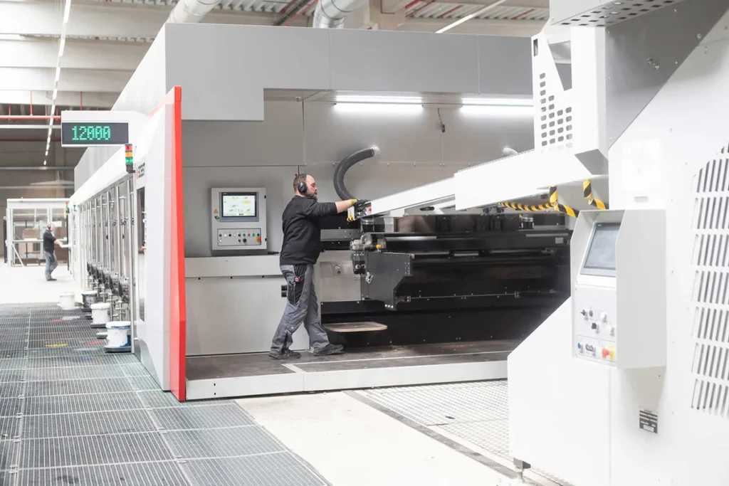 Mitarbeiter hebt einen großen Kartonzuschnitt an einer BOBST-Maschine.