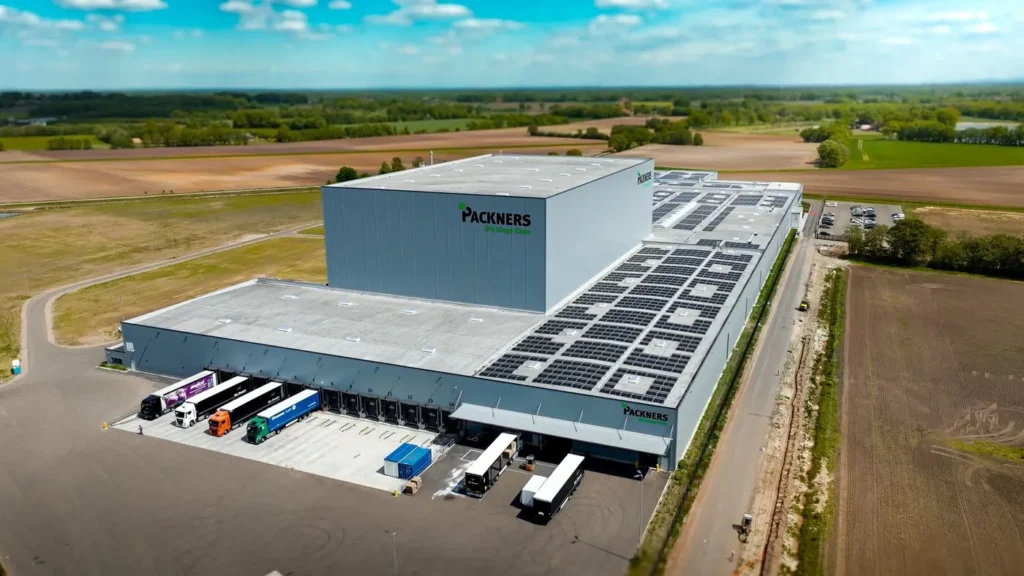 Luftaufnahme des neuen Packners Werks mit großflächigen Solaranlagen auf dem Dach und Lkw-Verladezonen im ländlichen Umfeld