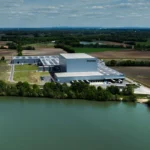 Weite Luftaufnahme des Packners-Werks am Wasser mit umliegenden Feldern.